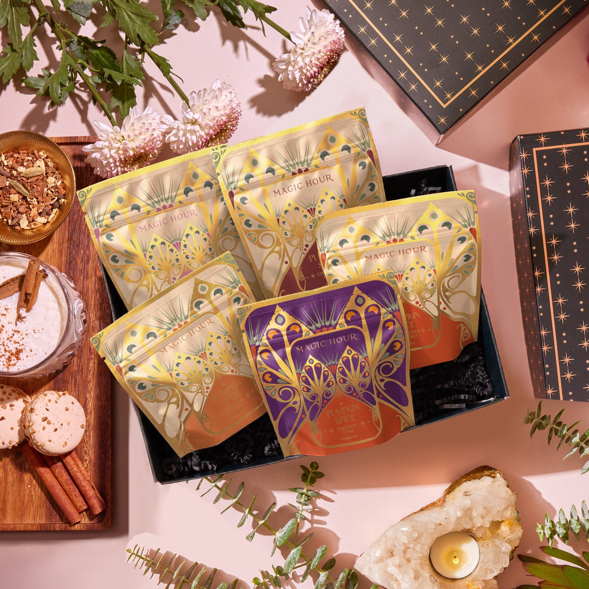 A Pumpkin Spice Abundance Sampler Bundle gift box containing five pouches with ornate floral illustrations, surrounded by a bowl of tea leaves, a spiced latte, two macarons, cinnamon sticks, fresh flowers, and a crystal candle holder.
