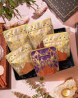 A Pumpkin Spice Abundance Sampler Bundle gift box containing five pouches with ornate floral illustrations, surrounded by a bowl of tea leaves, a spiced latte, two macarons, cinnamon sticks, fresh flowers, and a crystal candle holder.