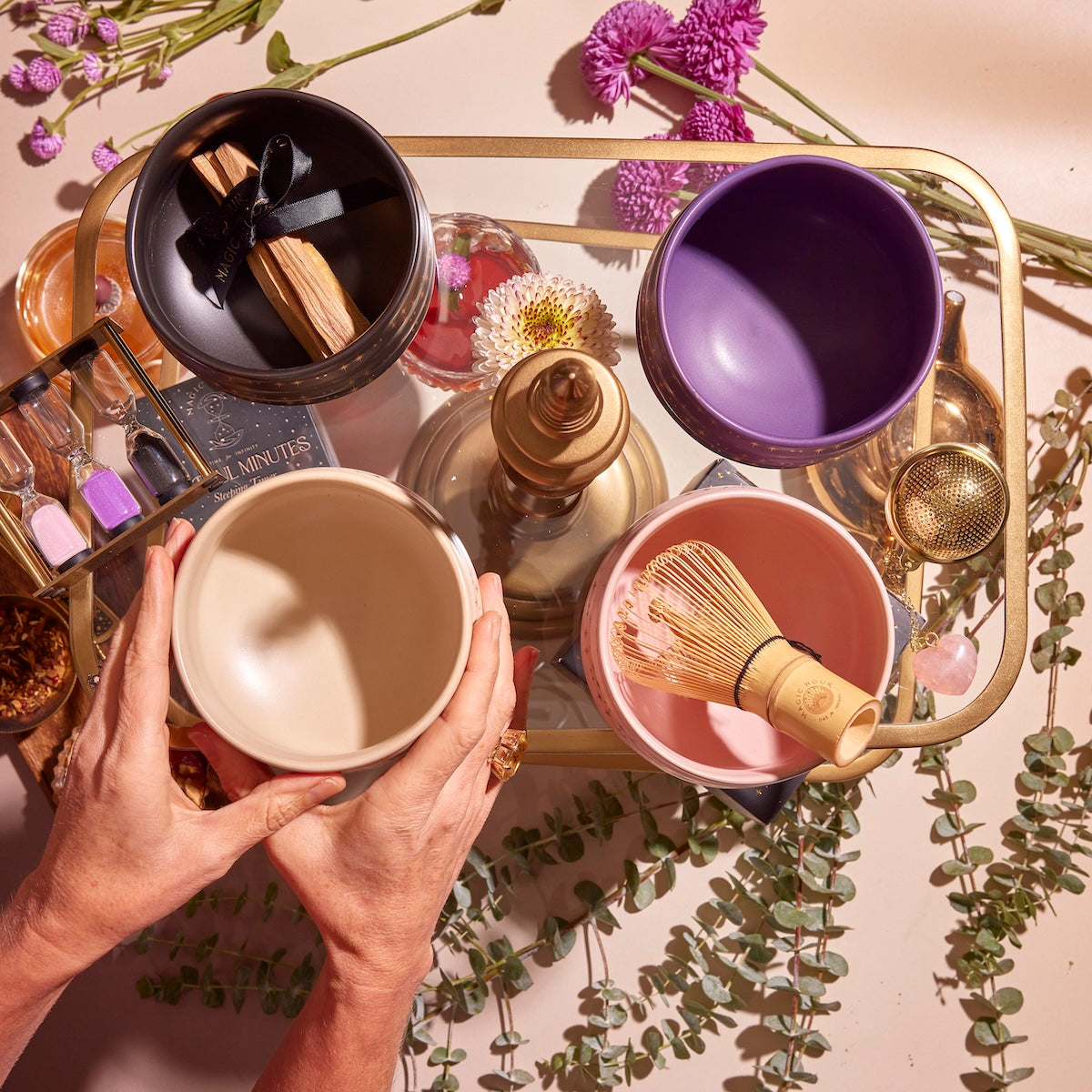 A top view of a person arranging four Matcha Bowls in beige, purple, and pink on a golden tray surrounded by scattered flowers and eucalyptus leaves. One bowl contains palo santo sticks with a black ribbon, while another holds a bamboo whisk, creating an elegant and serene scene.