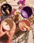 A top view of a person arranging four Matcha Bowls in beige, purple, and pink on a golden tray surrounded by scattered flowers and eucalyptus leaves. One bowl contains palo santo sticks with a black ribbon, while another holds a bamboo whisk, creating an elegant and serene scene.