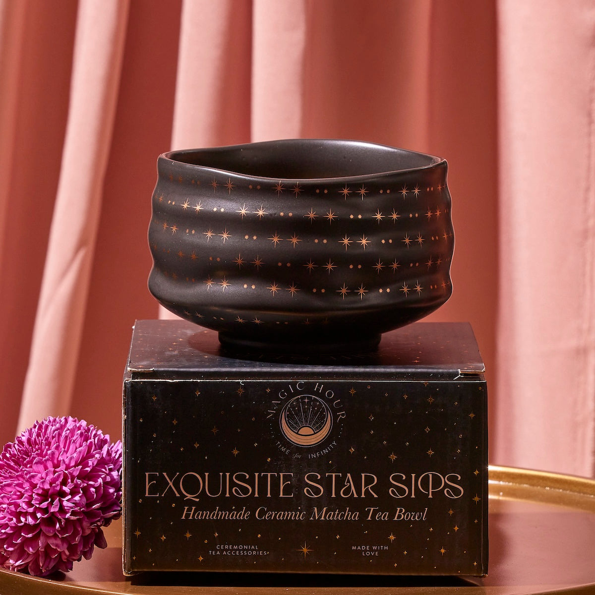 A dark, textured Matcha Bowl adorned with a star pattern rests on a black box labeled "Exquisite Star Sips: Handmade Ceramic Matcha Tea Bowl." Crafted using traditional methods and kiln firing, the background features soft pink drapes, and a purple chrysanthemum flower lies next to the box on a gold surface.