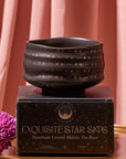 A dark, textured Matcha Bowl adorned with a star pattern rests on a black box labeled "Exquisite Star Sips: Handmade Ceramic Matcha Tea Bowl." Crafted using traditional methods and kiln firing, the background features soft pink drapes, and a purple chrysanthemum flower lies next to the box on a gold surface.