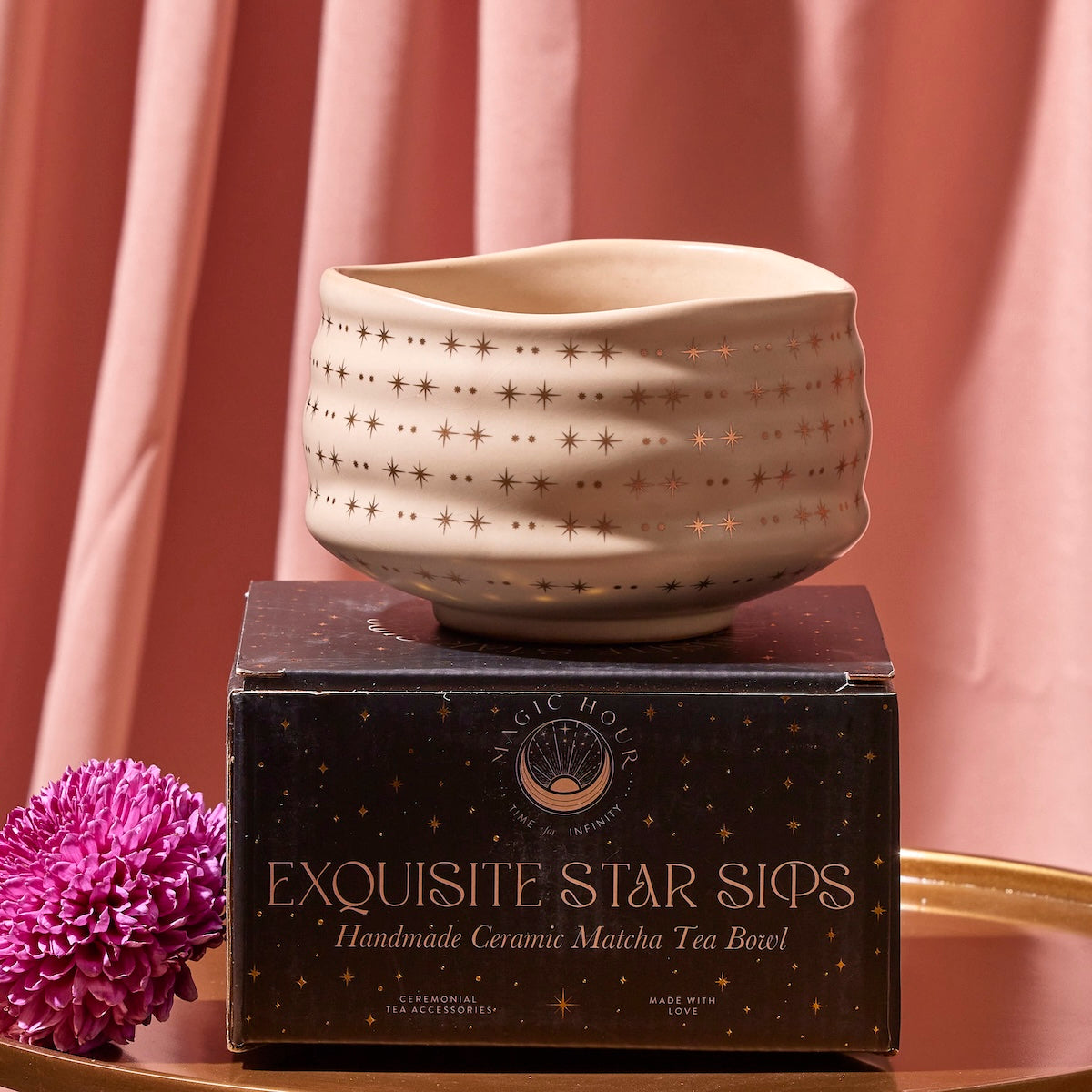 A Matcha Bowl featuring a starry design sits atop a black box labeled "Exquisite Star Sips." Crafted using traditional methods and kiln-fired, the bowl is displayed in front of a pink curtain, with a purple flower placed next to it.