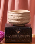 A Matcha Bowl featuring a starry design sits atop a black box labeled "Exquisite Star Sips." Crafted using traditional methods and kiln-fired, the bowl is displayed in front of a pink curtain, with a purple flower placed next to it.