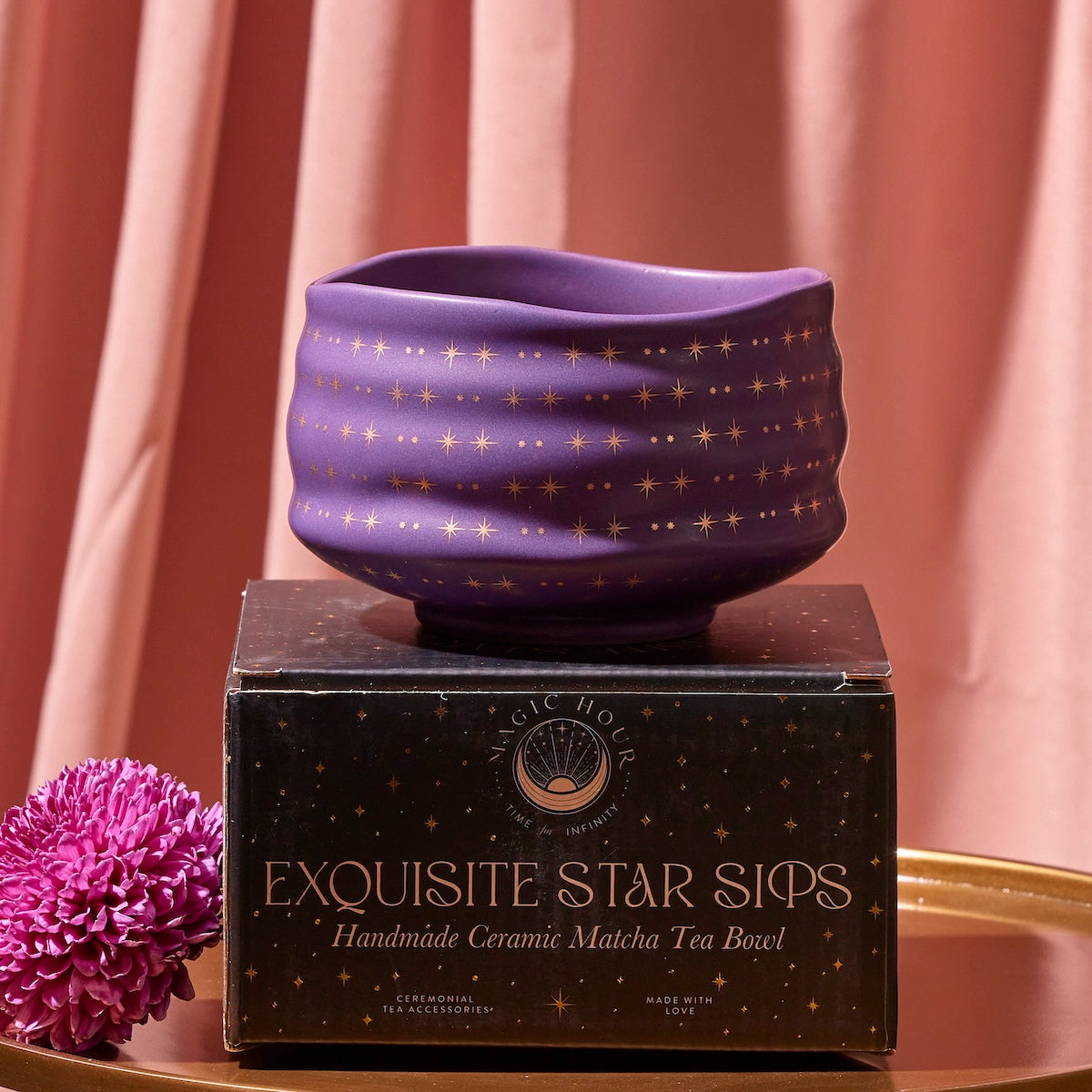 A handmade Matcha Bowl with a dark purple finish and gold star pattern, crafted by skilled artisans using traditional methods, sits atop a matching black and gold box labeled &quot;Exquisite Star Sips.&quot; A pink flower and pink draped curtain serve as a backdrop on a golden surface.