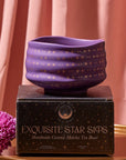 A handmade Matcha Bowl with a dark purple finish and gold star pattern, crafted by skilled artisans using traditional methods, sits atop a matching black and gold box labeled "Exquisite Star Sips." A pink flower and pink draped curtain serve as a backdrop on a golden surface.