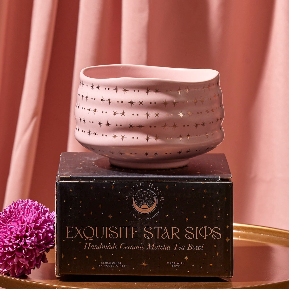 A pink Matcha Bowl with gold star patterns, crafted by skilled artisans using traditional methods, sits atop a black box labeled &quot;Exquisite Star Sips: Handmade Ceramic Matcha Tea Bowl.&quot; The background is adorned with matching pink fabric, and a purple chrysanthemum flower is placed to the left of the box.