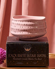 A pink Matcha Bowl with gold star patterns, crafted by skilled artisans using traditional methods, sits atop a black box labeled "Exquisite Star Sips: Handmade Ceramic Matcha Tea Bowl." The background is adorned with matching pink fabric, and a purple chrysanthemum flower is placed to the left of the box.