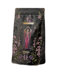 The Monthly Magic First Sips Tea Subscription Box includes a decorative tea package adorned with an illustration of a woman in a pink dress with flowing robes, holding two cups, reminiscent of Tarot archetypes. Surrounded by stars and purple flowers, the label reads “Magic Hour, The Queen of Cups,” providing an enchanting wellness tea experience.