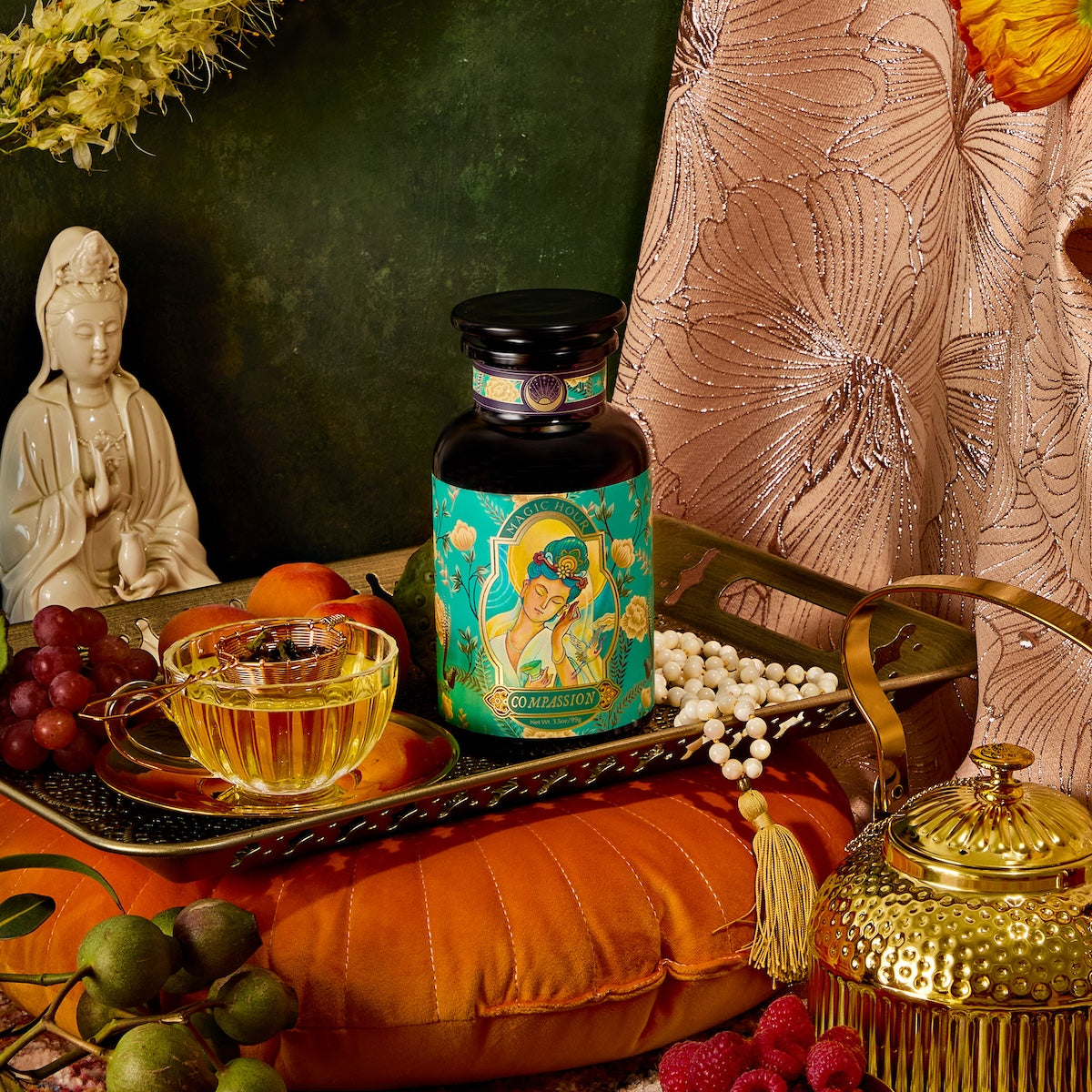 A tranquil scene features a Quan Yin statue, a &quot;Compassion&quot; jar, a glass teacup with saucer, a golden teapot, grapes, and berries arranged on an orange cushion. Draped fabric and flowers enhance the elegance.