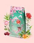 Introducing Queen of Kyoto: a vibrant tea package inspired by the Japanese Cherry Blossom Festival, adorned with pink cherry blossoms, a graceful crane, and elegant gold accents. The design is beautifully framed by roses, white flowers, cherries, and apricots against a soft pink background.