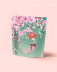 A pastel green tea pouch with pink cherry blossoms, white flying birds, and gold accents evokes the Japanese Cherry Blossom Festival. The label reads “Queen of Kyoto” and hints at delicate Sencha Green Tea within.