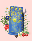 A blue and yellow tea package labeled "Queen of Ukraine: Spring Blossom Tea" stands amid colorful flowers, strawberries, and vanilla beans on a light pink background. The ornate packaging, adorned with a sun and floral designs, hints at the delightful tea blend within.