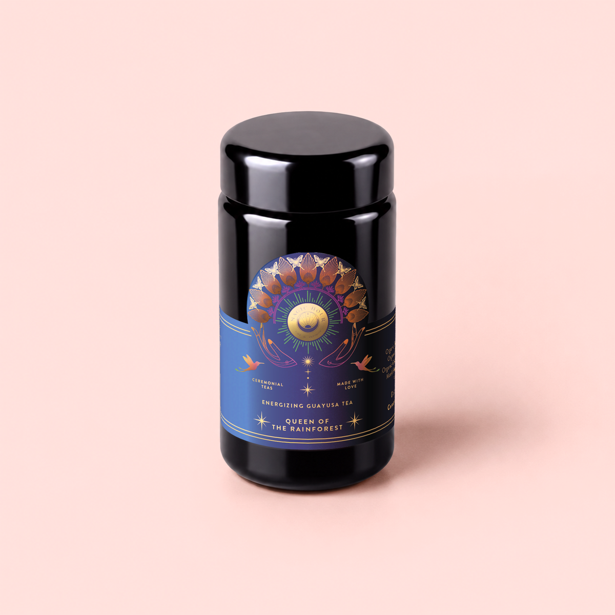 A black tea canister with a colorful sun and moon motif on a blue label reading “Queen of the Rainforest : Cacao-Berry Tea” sits against a pale pink background, perfectly capturing the vibrant rainforest energy found in every sip of this unique cacao-berry infused Guayusa tea.