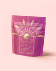 A pink resealable pouch labeled "Queen of the Tropics: Rosé all Day Alternative Tea" features gold botanical and sunburst designs—an elegant herbal rose tea with Garcinia Cambogia, offering a refined alternative to Rosé wine.