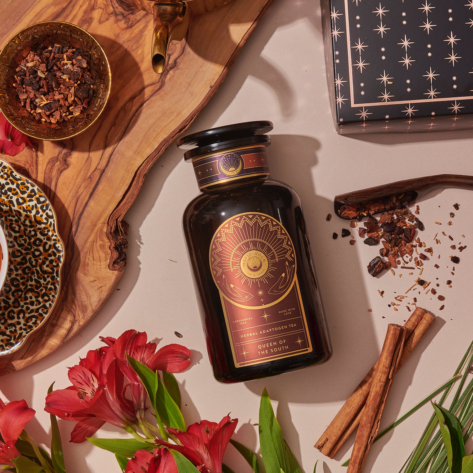 A dark glass bottle of Queen of the South Cocoa Detox Tea rests on a beige surface with red flowers, cinnamon sticks, dried herbs in a brass bowl, a patterned plate, and a black star box—ideal for steeping this adaptogenic tea blend.