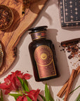 A dark glass bottle of Queen of the South Cocoa Detox Tea rests on a beige surface with red flowers, cinnamon sticks, dried herbs in a brass bowl, a patterned plate, and a black star box—ideal for steeping this adaptogenic tea blend.