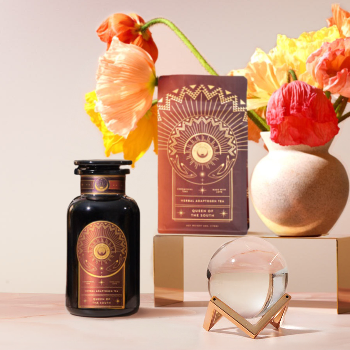 The Queen of the South Cocoa Detox Starter Kit—a black jar and box with gold sunburst motifs—sits by a round glass orb, while orange and yellow poppies brighten the background in a beige vase.