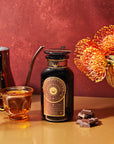 A glass teacup filled with caffeine-free, adaptogenic tea sits next to a decorative bottle labeled "Queen of the South: Delicious Cocoa Detox Tea." Nearby, a metallic kettle and a golden vase with orange flowers are displayed alongside small pieces of chocolate on a red background.