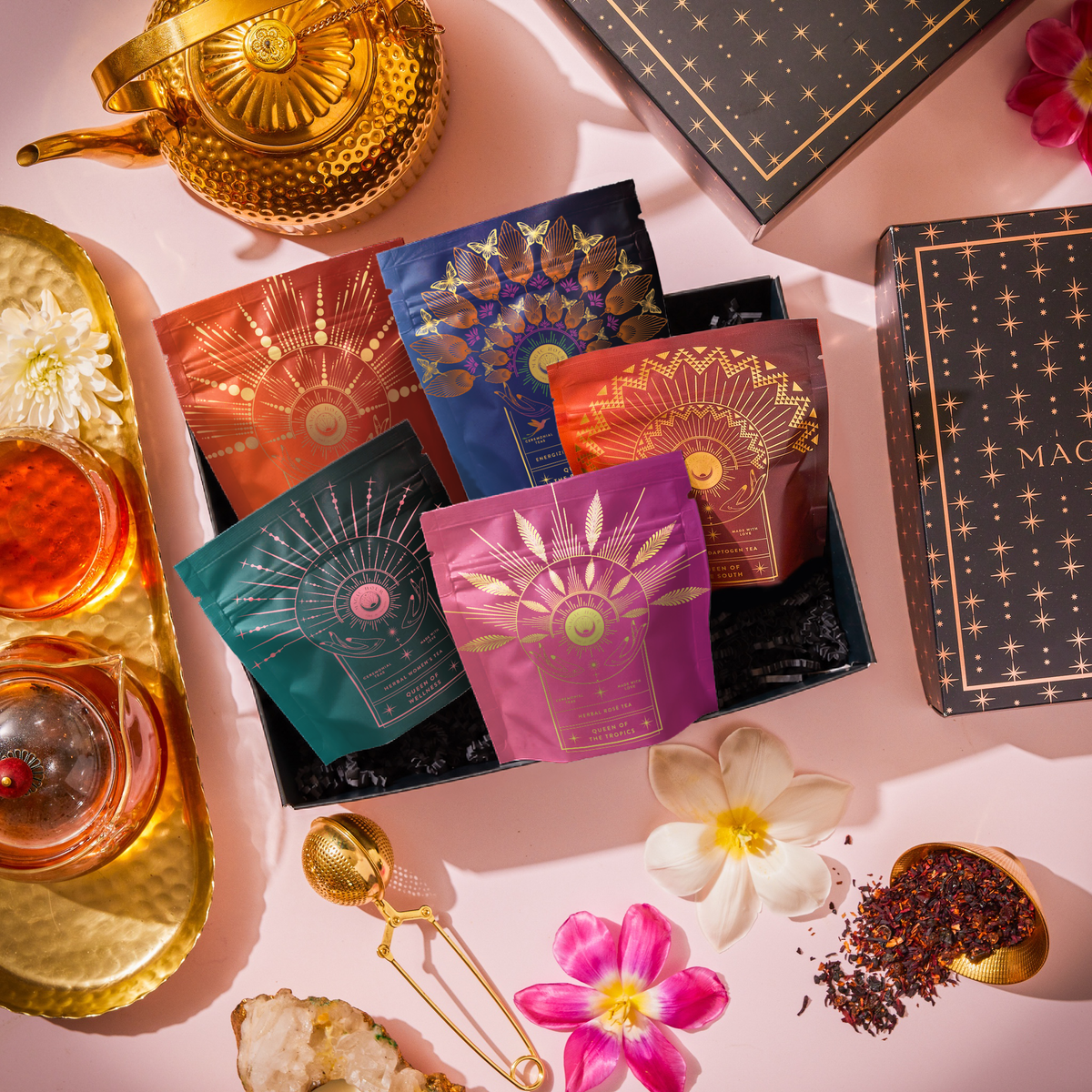 The Queen’s Sampler Set of five colorful tea pouches decorated with gold celestial designs, arranged on a surface with a gold teapot, glass teacups, and loose wellness teas.