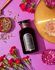 A black bottle labeled "Raspberry Earl White Tea" lies on a vibrant purple background, accompanied by a gold plate, scattered white tea leaves rich in antioxidants, red flowers, a small dish of raspberries, and a pink-lit candle in a rose-shaped holder.