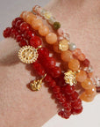 Wrist adorned with the **Red Carnelian Mandala Bracelet**, featuring multiple beaded strands in vibrant shades of red, orange, and translucent white. This healing jewelry piece also boasts gold decorative charms, including a compass, a horn, and a leaf. The beads come in various sizes, creating a colorful and layered look.
