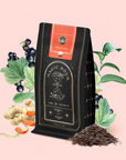 A bag of Magic Hour adaptogenic wellness tea labeled "Rise & Shine Black Tea" is surrounded by blackcurrants, ginger, leafy branches, and a small pile of loose tea leaves on a light pink background.