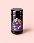A canister of Romance in Ravello - Chocolate-Passionfruit-Rose-Cream White Tea sits on a pale pink background, its purple floral label adorned with passion flowers and fruit, evoking inspiration from the Amalfi Coast.