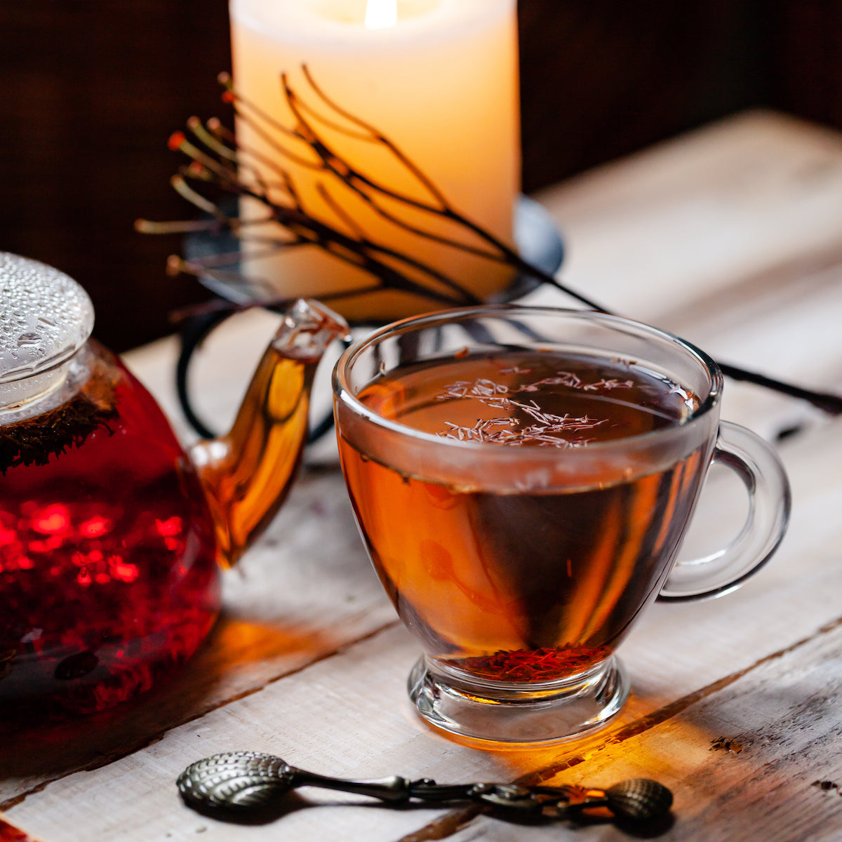 A glass cup of tea sits on a wooden table next to a glass teapot filled with tea. Behind the teapot, a lit candle on an iron stand creates a warm, cozy atmosphere. Two ornate silver teaspoons lie in the foreground.