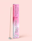 A box of Natural Incense : Rose Crystal, labeled “Love,” stands upright on a pink background beside a lit sandalwood incense stick in a small round holder.