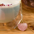 A clear glass cup containing a frothy beverage sits on a wooden surface. The drink is adorned with dried rose petals, and a chain with the Rose Quartz Heart Tea Strainer, featuring a pink heart-shaped pendant, dangles from the cup.