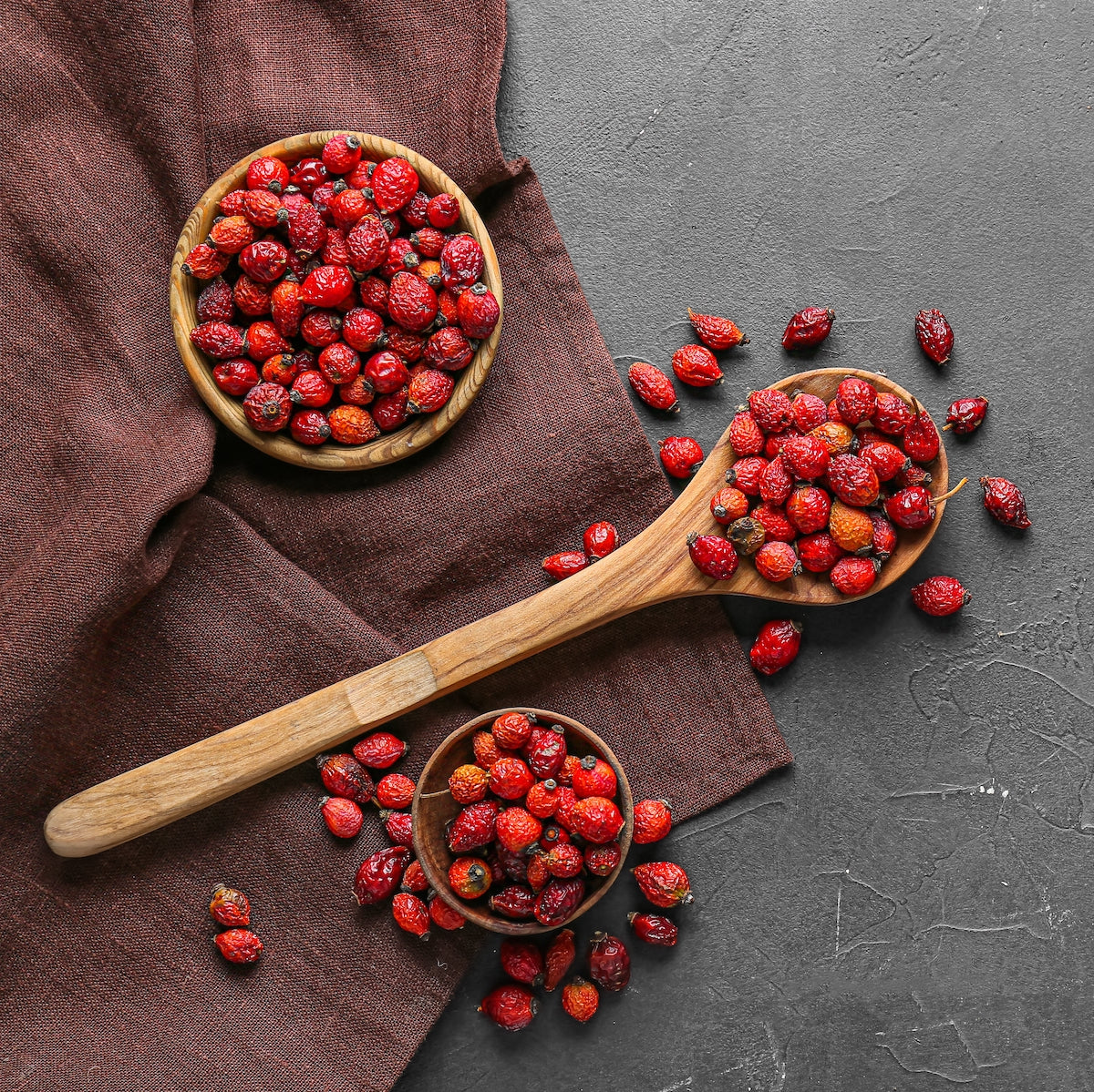 Top view of dried rosehips in small wooden bowls and on a wooden spoon over a brown cloth and gray textured surface. The red berries are scattered around, adding a vibrant contrast to the background.