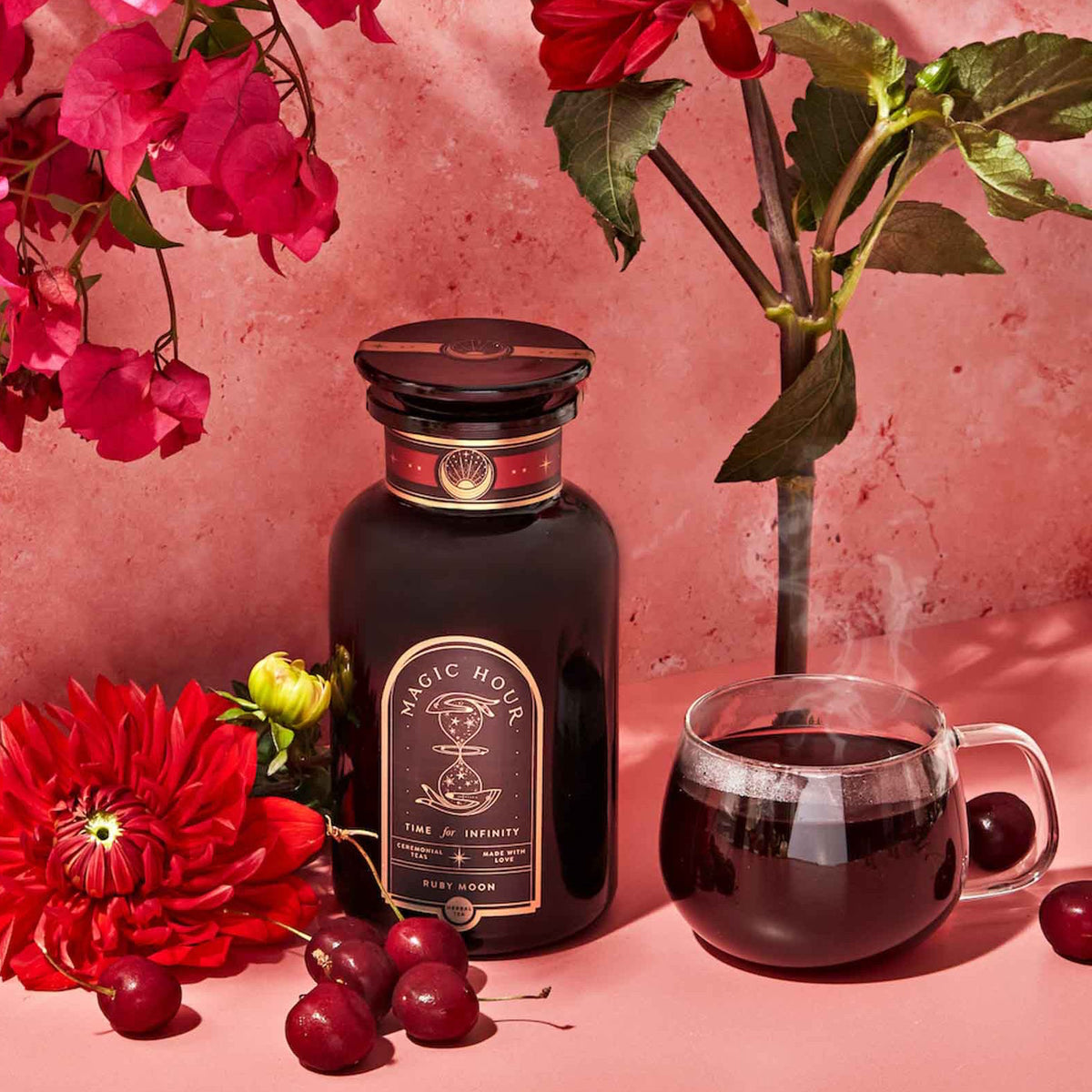 A dark apothecary jar of Best Sellers of Magic Hour Trio Box labeled “Ruby Moon” sits next to a glass mug of steaming dark tea, surrounded by red flowers and cherries.