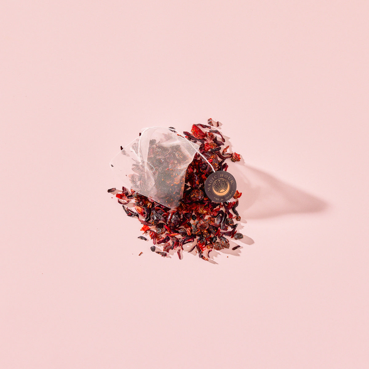 A pyramid tea bag of Tea for Manifesting: Ruby Moon: Hibiscus, Elderberry &amp; Passionfruit Herbal Tea resting on a pile of organic elderberry tea, loose-leaf dried hibiscus, and fruit pieces.