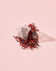 A pyramid tea bag of Tea for Manifesting: Ruby Moon: Hibiscus, Elderberry & Passionfruit Herbal Tea resting on a pile of organic elderberry tea, loose-leaf dried hibiscus, and fruit pieces.