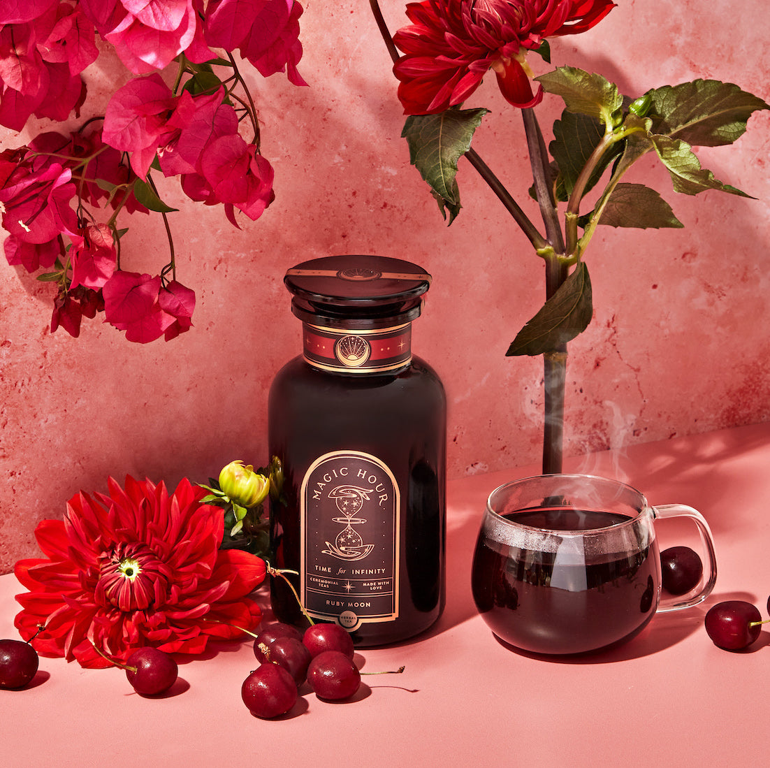 A still life arrangement featuring a glass bottle labeled "Magic Hour" surrounded by vibrant red flowers, cherries, and a steaming glass cup filled with a dark liquid. The background is a soft pink wall adorned with hanging red blooms.
