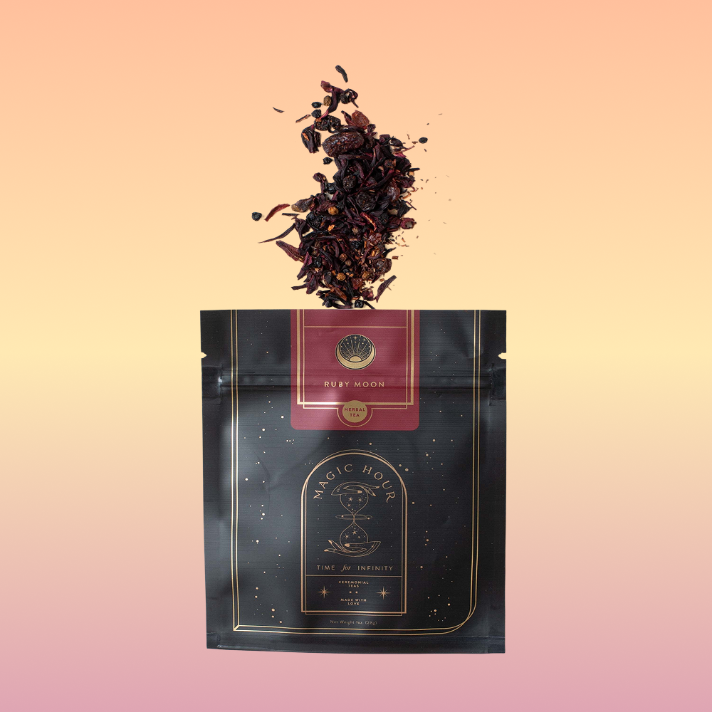 A black and gold pouch labeled "Ruby Moon™ : Hibiscus Elderberry Tea" rests against a white background. The pouch, containing organic loose leaf tea by Club Magic Hour, features galaxy-themed designs. Beside it lies a small arrangement of purple and white orchids.