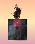 A black and gold pouch labeled "Ruby Moon™ : Hibiscus Elderberry Tea" rests against a white background. The pouch, containing organic loose leaf tea by Club Magic Hour, features galaxy-themed designs. Beside it lies a small arrangement of purple and white orchids.