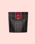 A black and gold resealable pouch labeled "Magic Hour" with a red top that says "Ruby Moon™ Herbal Tea Bags." The elegant celestial design highlights this caffeine-free hibiscus tea against a plain light gray background.