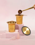 A decorative gold cup holds organic loose leaf tea, with a hand using a gold scoop to add more. Another identical gold cup and a lid sit nearby on two white platforms. Two small pink crystals are placed in the foreground against a soft pink background, creating the perfect Magic Hour Steep The Stars - Artisan Tea Strainer setting.