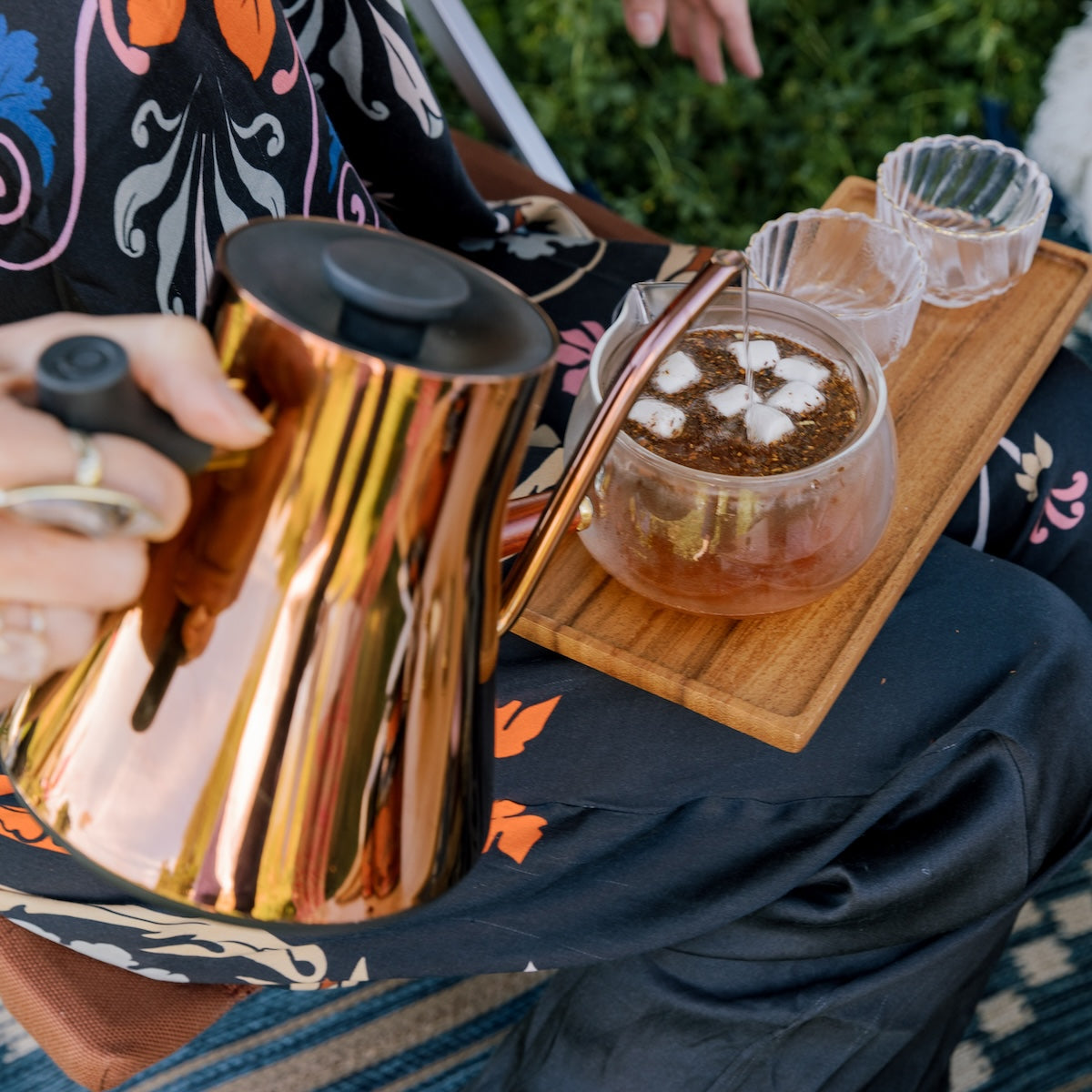 A copper tea kettle pouring hot water into a glass teapot filled with S&#39;mores Herbal Dessert Loose Leaf Wellness Tea on a Wooden tea tray.