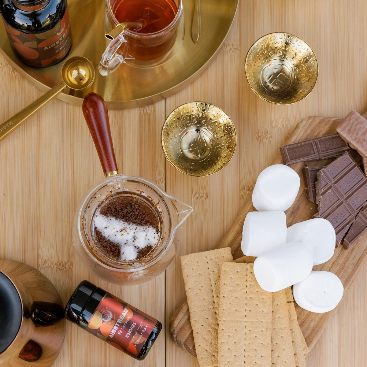 A top-down view of a wooden table set for s&#39;mores preparation. Ingredients include marshmallows, graham crackers, and chocolate bars on a wooden board. A cup of S&#39;mores Tea by Magic Hour, tea-making utensils with loose leaf tea, a jar of spice, and two ornate golden cups are also present.