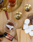 A top-down view of a wooden table set for s'mores preparation. Ingredients include marshmallows, graham crackers, and chocolate bars on a wooden board. A cup of S'mores Tea by Magic Hour, tea-making utensils with loose leaf tea, a jar of spice, and two ornate golden cups are also present.