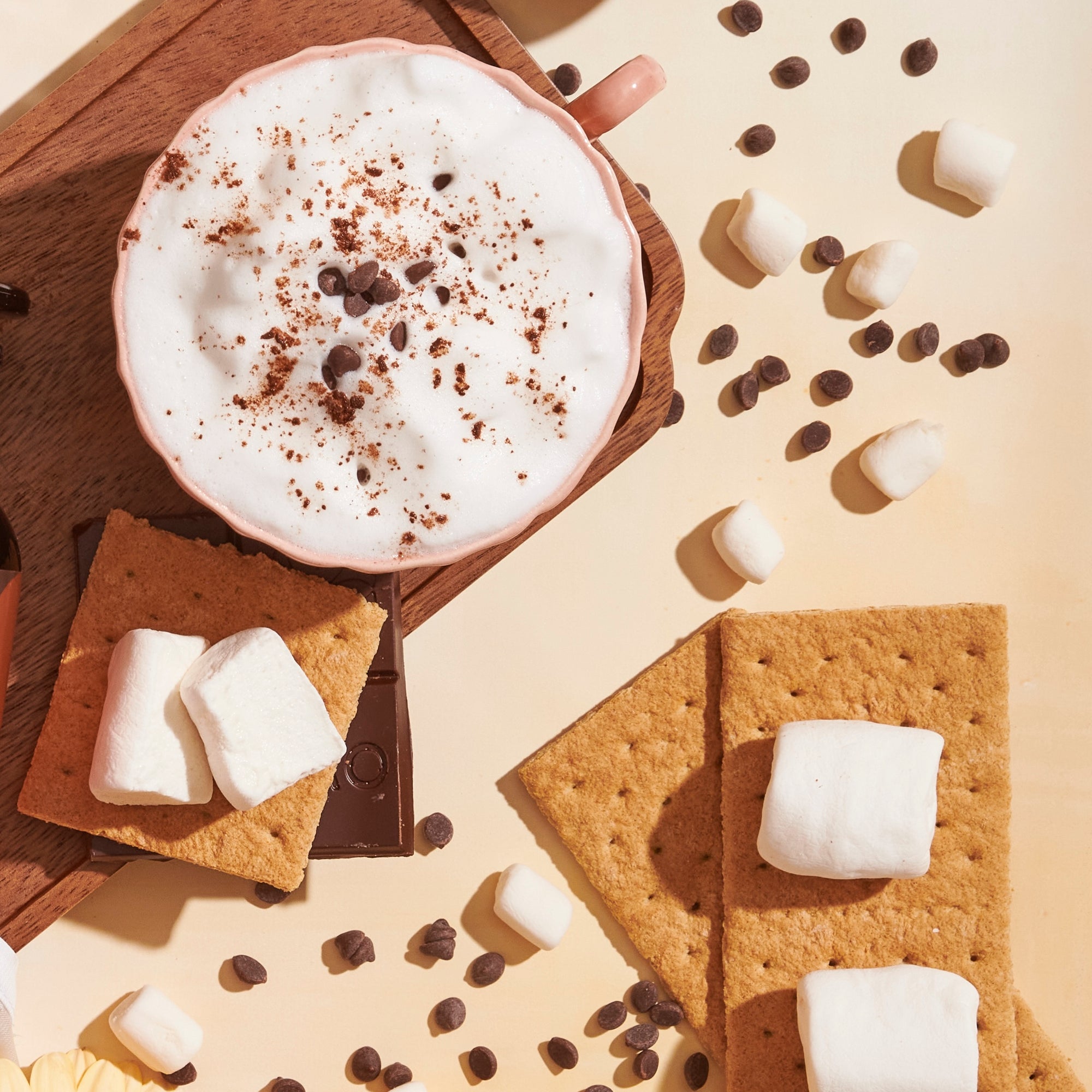 a S&#39;mores tea latte with foamed milk, chocolate and cinnamon and chocolate chips sprinkled on the table with graham crackers, chocolate, and marshmallows,