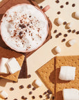 a S'mores tea latte with foamed milk, chocolate and cinnamon and chocolate chips sprinkled on the table with graham crackers, chocolate, and marshmallows,