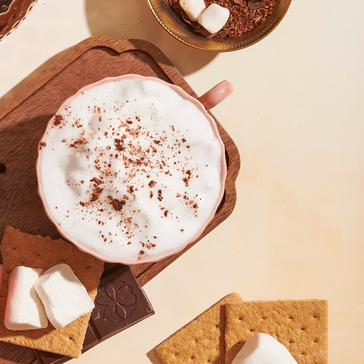 A cup of frothy cappuccino sprinkled with cocoa powder sits on a wooden tray. Surrounding the tray are graham crackers, a small piece of chocolate with a design on it, and marshmallows. Embrace the ultimate summer tea experience with the Hot Girl Summer Tea Box (Put it on ICE!) while savoring this delightful setup.