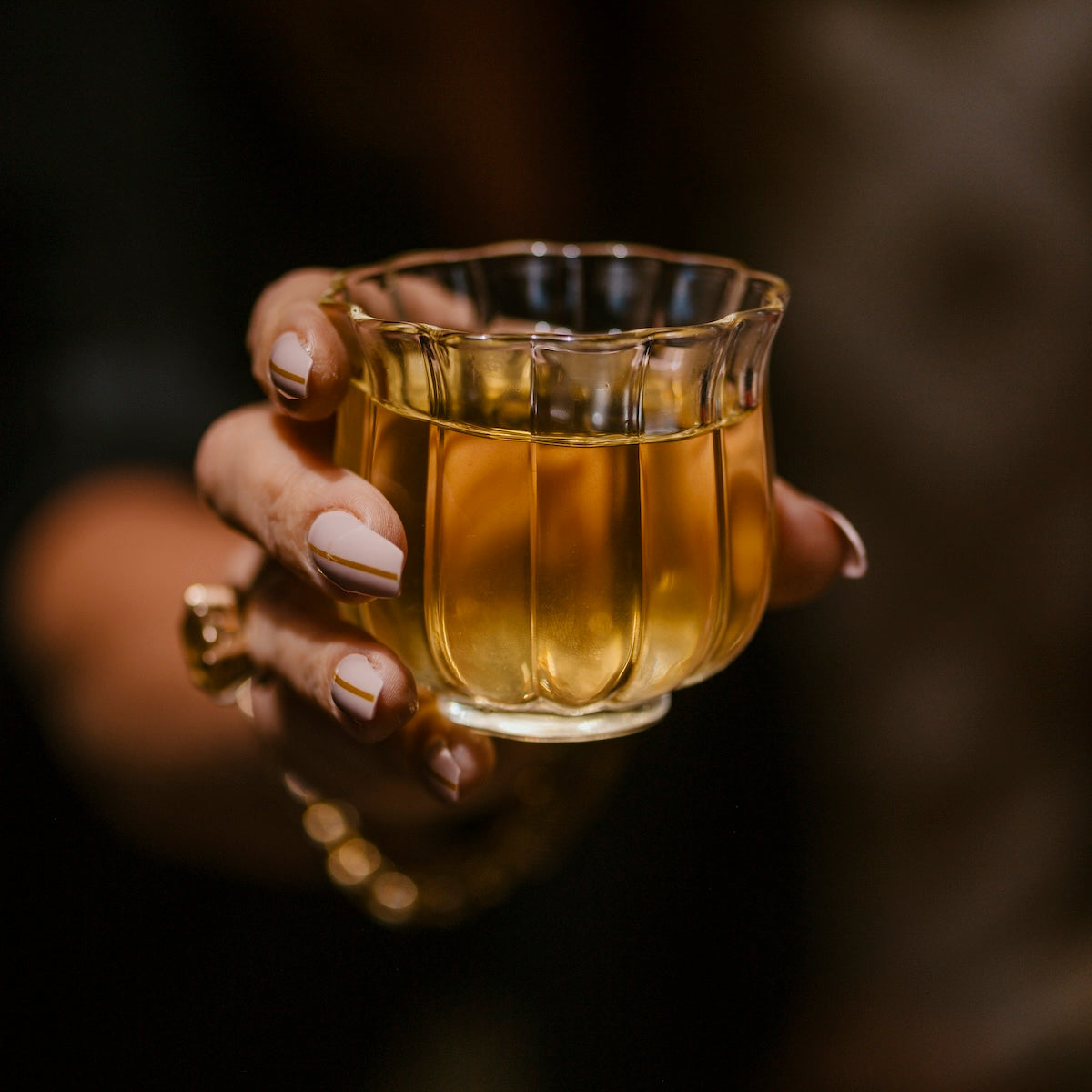 A hand with beige manicure and a gold ring holds an intricately designed, translucent glass filled with a golden liquid, possibly April 2024 Harvest - Grand Cru First Flush Goomtee Estate Darjeeling FTGFOP1. The background is dark, making the glass and exquisite tea from Magic Hour the focal point of the image.