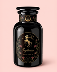 A black glass perfume bottle showcases gold Sagittarius art, flowers, and stars on its label. The text reads “Sagittarius” and “Tea of Good Fortune & Abundance.” The enchanting scene suggests the invigorating aroma of pomegranate ginger tea against a soft pink background.