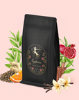 A black bag labeled "Sagittarius Tea of Good Fortune & Abundance" is surrounded by leaves, pomegranate, orange slice, ginger, vanilla flower, cinnamon sticks, and tea leaves—evoking a mood-boosting pomegranate ginger tea.