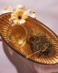 A glass teacup filled with Magic Hour's April 2024 Harvest - Grand Cru First Flush Organic "Spring Wonder" Samabeong Estate Darjeeling STGFOP1 on a golden oval tray, next to a small heap of loose organic tea leaves and a golden spoon. The tray is adorned with a single yellow gerbera daisy and white rose petals, all set against a soft pink background.